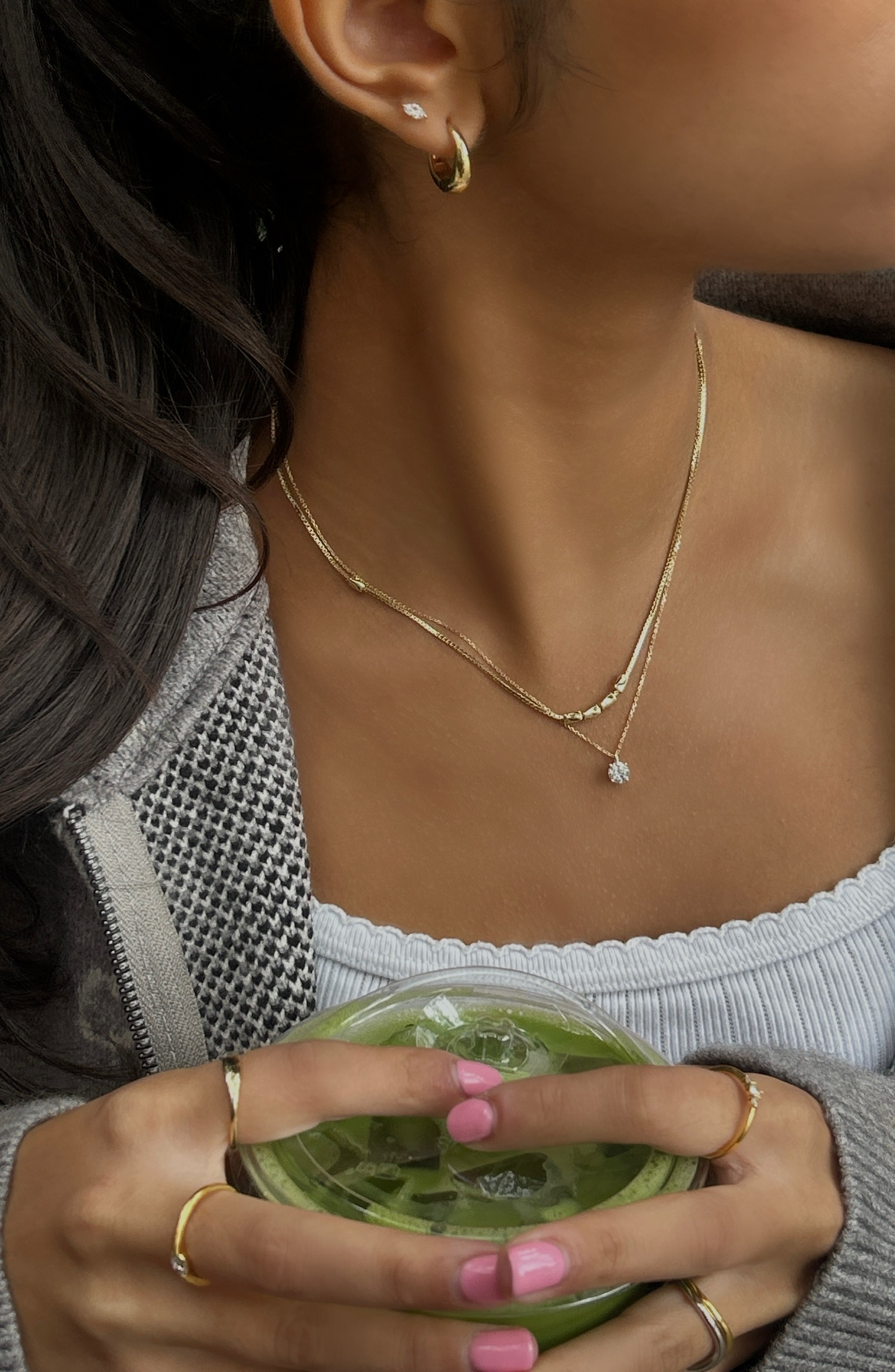Close-up of earrings, layered necklaces and rings worn with a checked jacket while holding a green drink